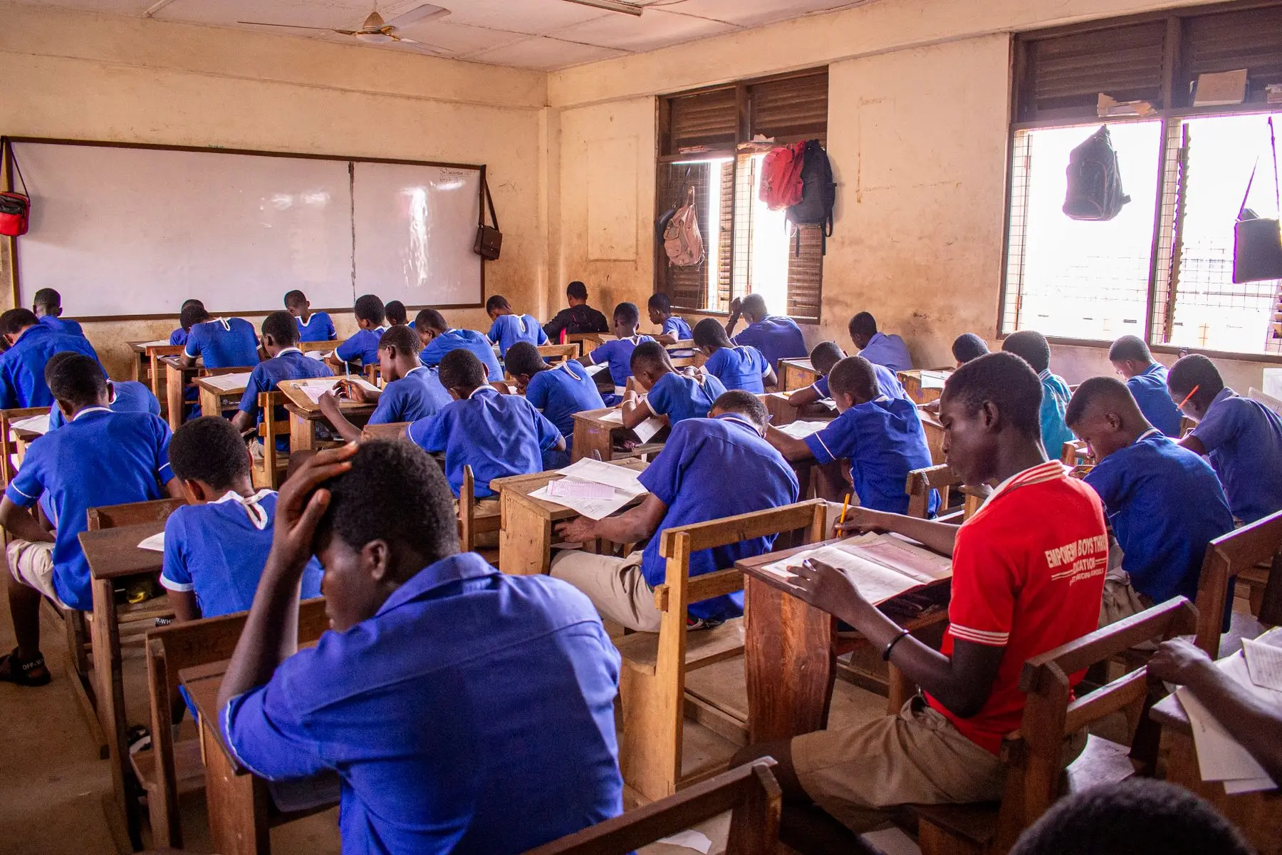 Learners sitting an examination at wooden desks in a full East African classroom